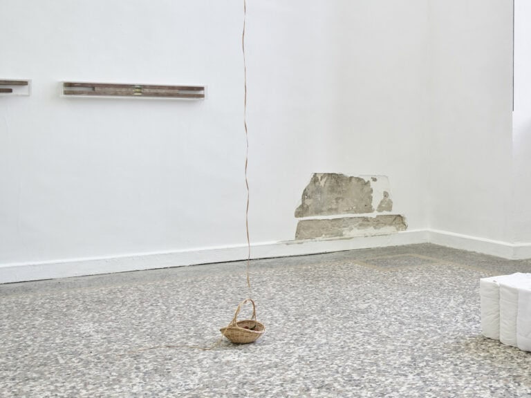 Foto: Ansicht eines Raumes mit weißen Wänden und einem mehrfarbigen Steinboden. An den Wänden und auf dem Boden befindet sich Objekte. An einem kleinen, bodennahmen Teil der Wand ist die Farbe abgeschlagen und die damunterliegende Schicht des Gemäuers ist sichtbar. Auf dem Boden steht ein Korb ,von dem ausgehend eine Schnur aus dem Bildraum heraus zur Decke reicht.