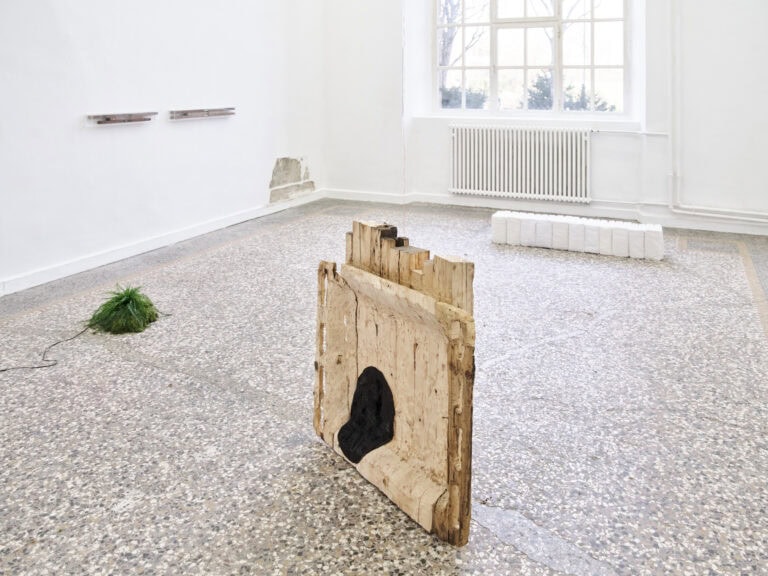 Foto: Ansicht eines Raumes mit weißen Wänden, einem Fenster und einem mehrfarbigen Steinboden. An den Wänden und auf dem Boden befindet sich Objekte. An einem kleinen, bodennahmen Teil der Wand ist die Farbe abgeschlagen und die damunterliegende Schicht des Gemäuers ist sichtbar.