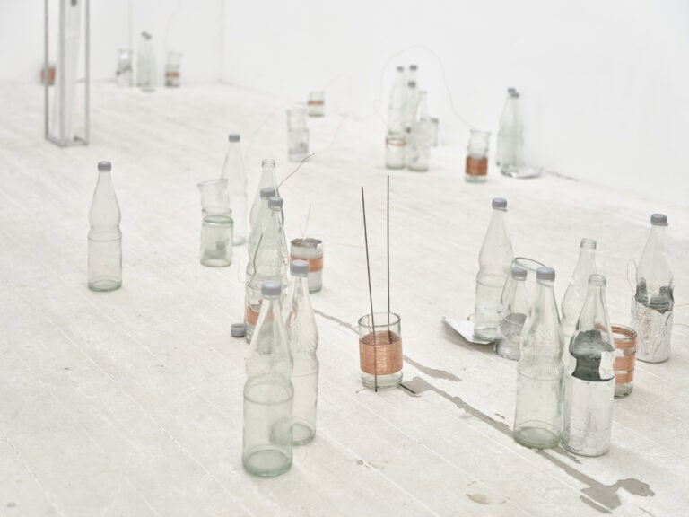 Foto: Nahaufnahme. Eine Vielzahl an leeren Flaschen und Gläsern auf einem gräulichen Boden. Um manche der Objekte ist ein kupferfarbener Draht gewickelt. Manche der Flaschen sind geöffnet, andere geschlossen.