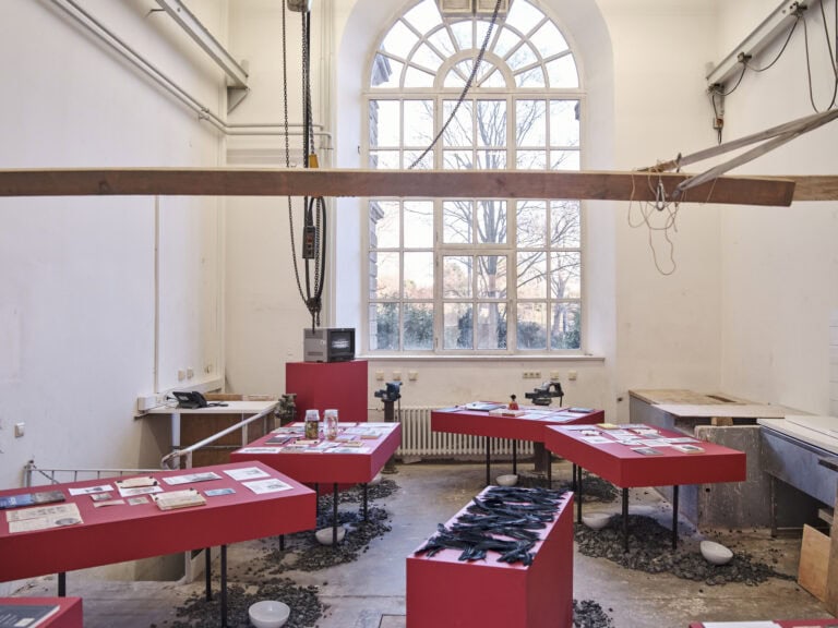 Foto: Ansicht eines Raumes mit hellen Wänden, einem Bogenfenster und gräulichem Boden. Auf dem Boden des Raumes befinden sich graue Steine und weiße, leere Schüsseln. Im Raum stehen Tische mit roten, tiefen Tischplatten und rote Podeste. Auf einem der Podest liegen schwarze Federn, auf dem anderen steht ein quadratischer Fernsehener. Auf den Tischen liegen verschiedene Objekte, Zettel, Bücher und gefüllte Gläser. Oben im Bild ist ein Balken zu erkennen, der in Spanngurten im Raum schwebt.