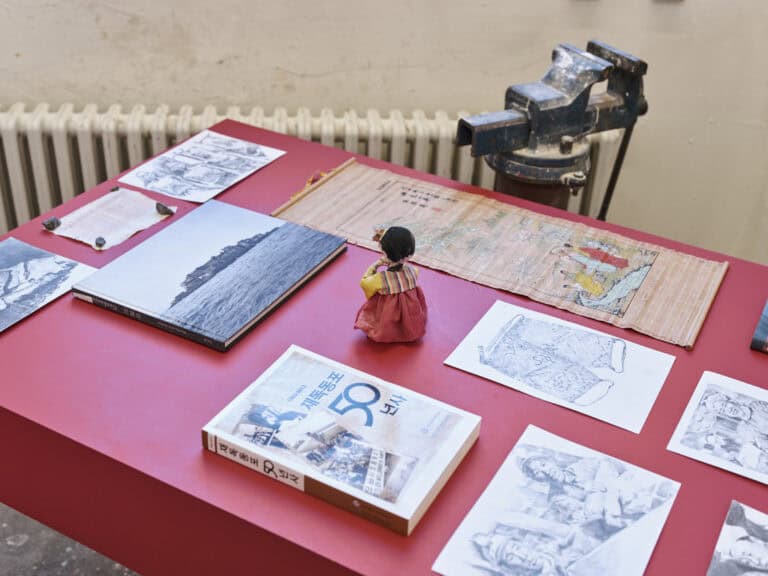 Foto: Nahansicht von Objekten, Büchern und Zetteln auf einer roten Oberfläche. Eines der Bücher hat koranische Buchstaben auf dem Umschlag.