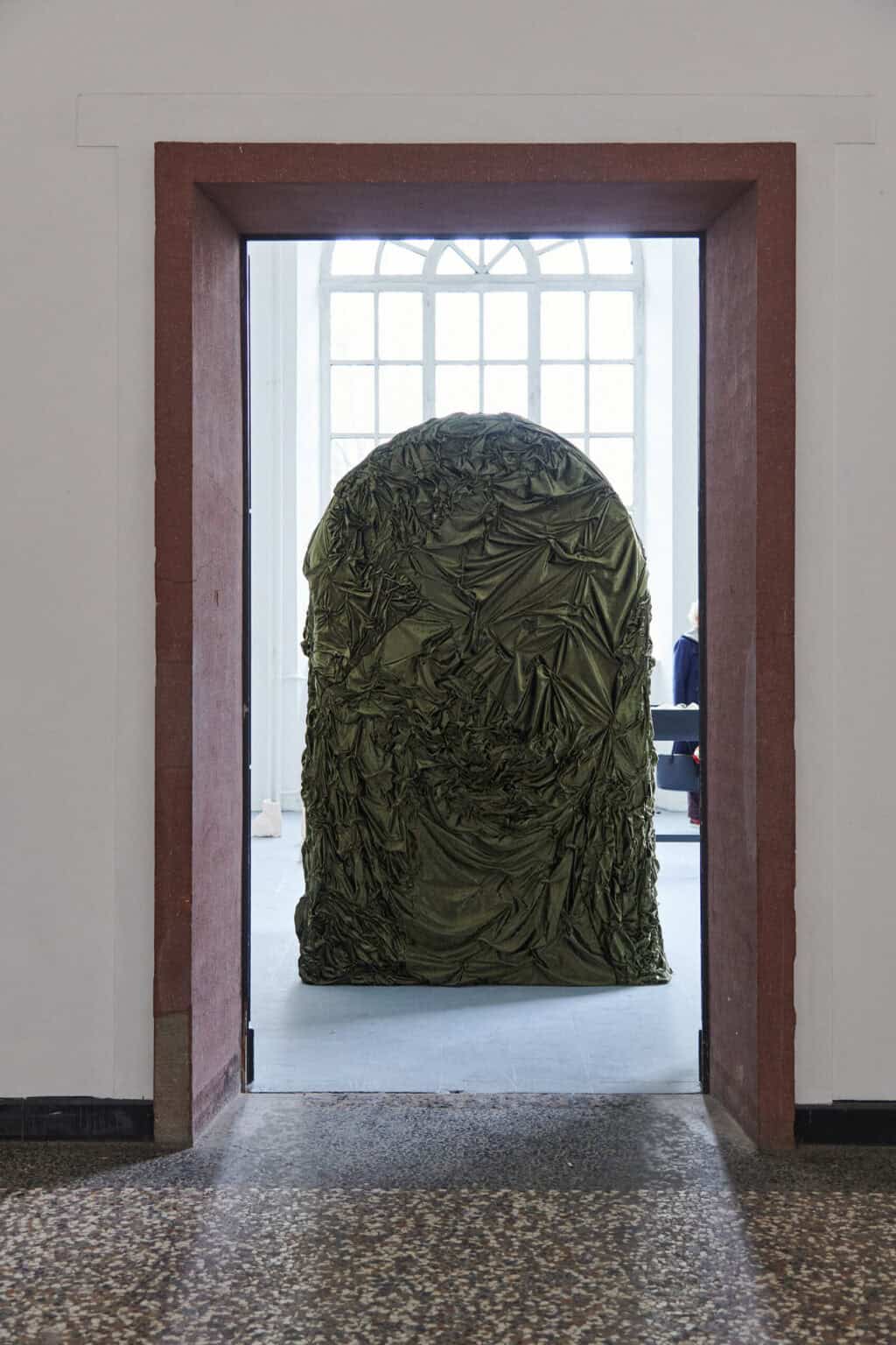 Foto: Ansicht eines massiven, hohen Türrahmens mit geöffneter Tür. Im Durchblick auf den hinterliegenden Raum steht ein großes, flächiges Objekt.