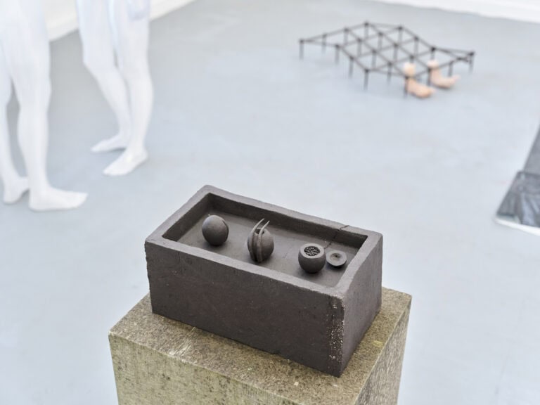 Foto: Nahaufnahme eines braunen Objektes auf einem braun-gelblichen Sockel. Im Hintergrund sind unscharf weitere Objekte und ein grauer Boden zu sehen.