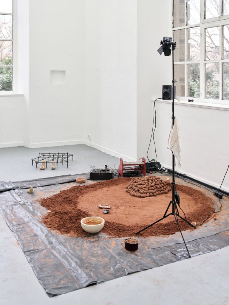 Foto: Ansicht eines Raumes mit weißen Wänden, zwei Fenstern und grauem Boden. In der Wand befindet sich eine rechteckige Einbuchtung. Auf dem Boden liegen Objekte, darunter auch eine schwarze Folie, auf der sich unterschiedliche Objekte, darunter erdiges Material und Technik, befinden. Auf der Fensterbank steht ein Lautsprecher, der mit der Technik verbunden ist.