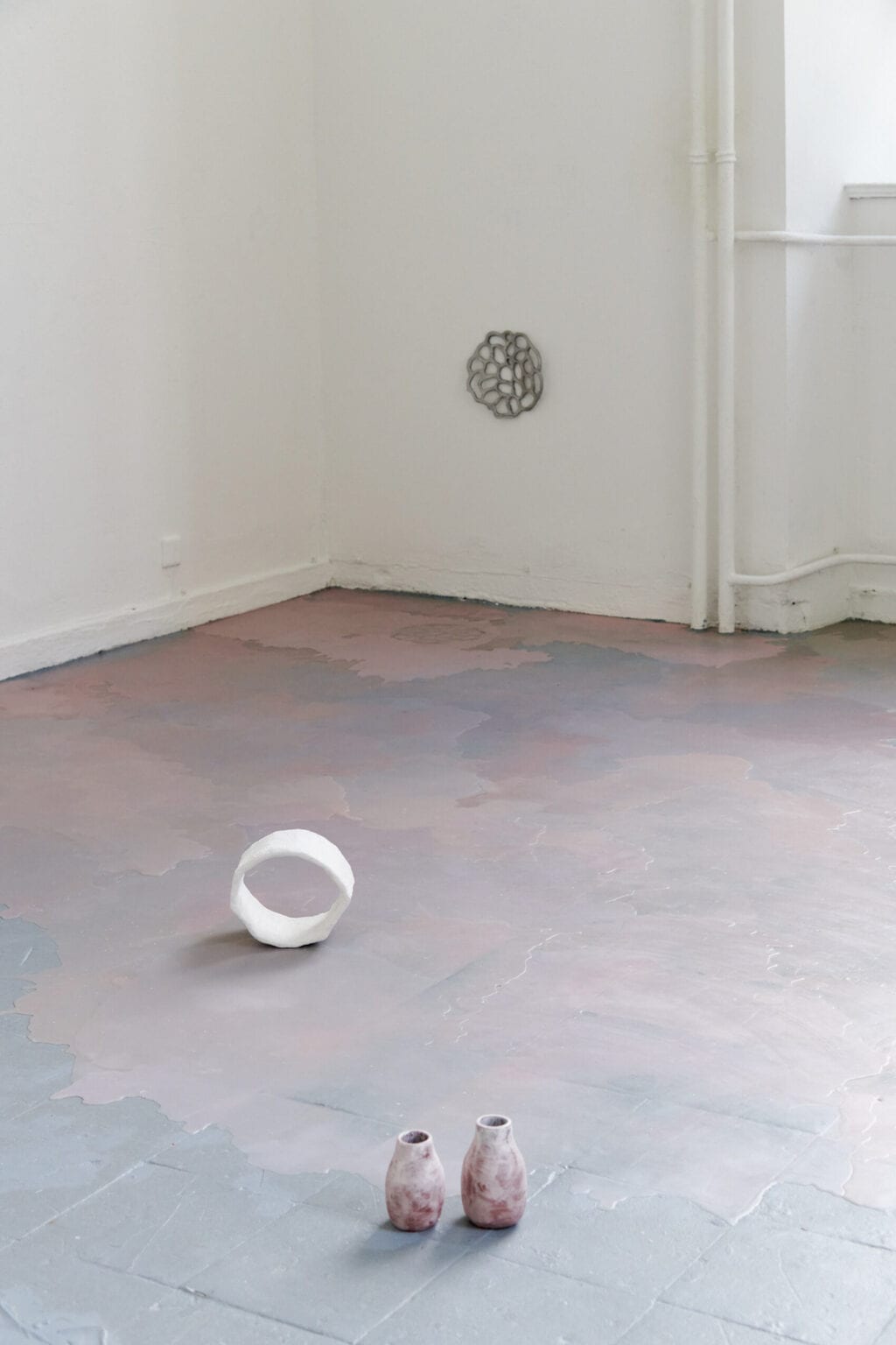 Foto: Ein Raum mit weißen Wänden, grauem Boden und einem Fenster. Auf dem Boden und an der Wand befinden sich Objekte. Teile des Bodens sind mit einer matten, rosafarbenen Masse bedeckt, die unebene, pfützenartige Konturen hat.