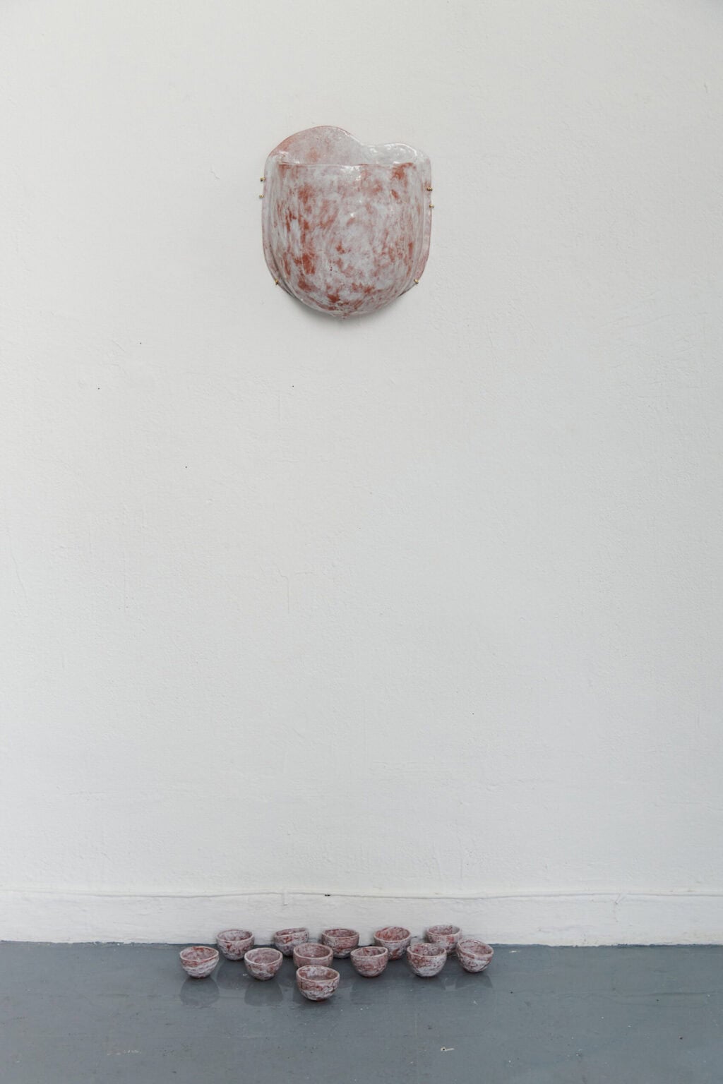 Foto: Ein Objekt an weißer Wand und wandnah eine Gruppe schalenförmiger Objekte auf dem grauen Boden.