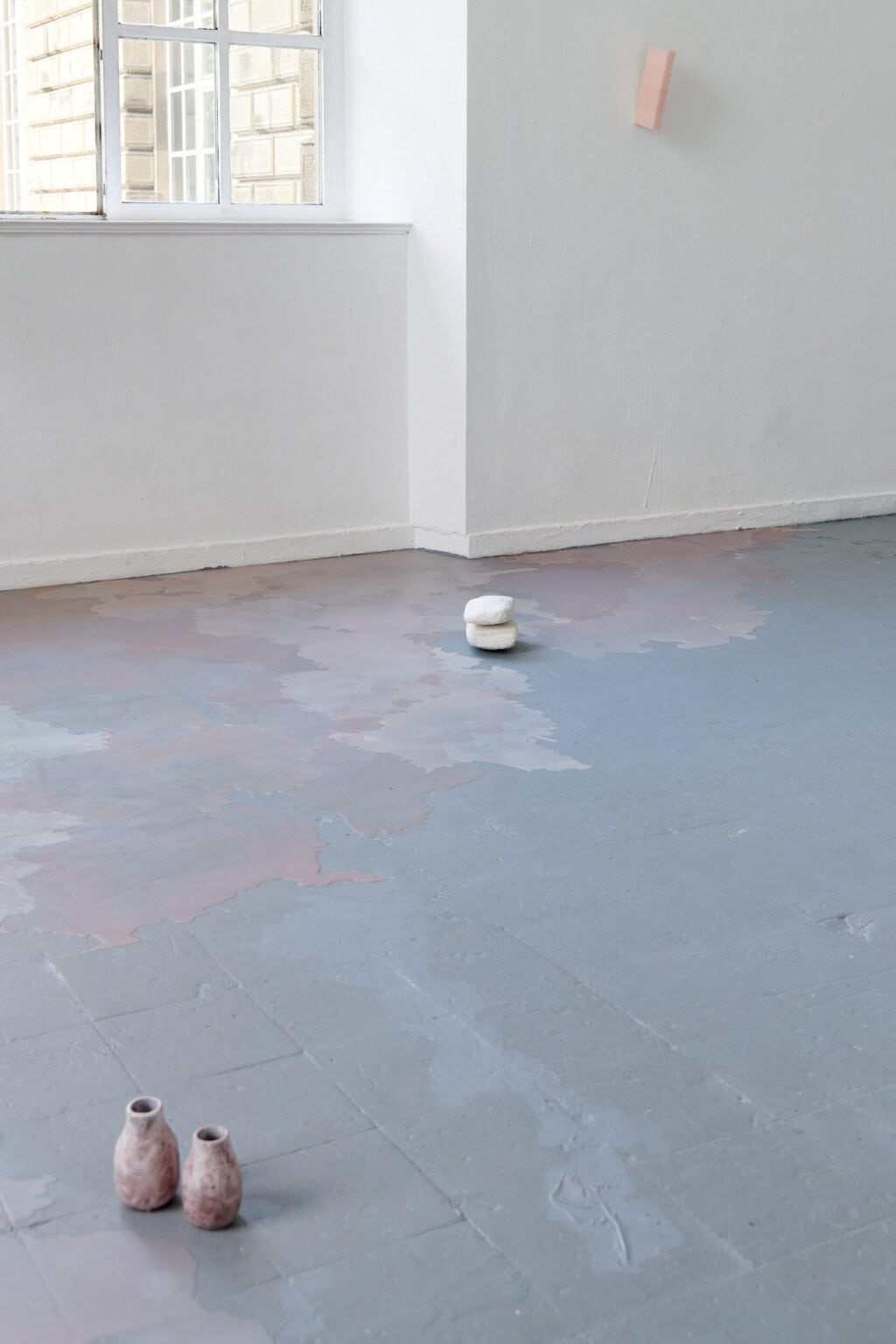 Foto: Ein Raum mit weißen Wänden, grauem Boden und einem Fenster. Auf dem Boden und an der Wand befinden sich Objekte. Teile des Bodens sind mit einer matten, rosafarbenen Masse bedeckt, die unebene, pfützenartige Konturen hat.