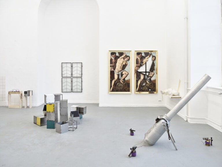 Foto: Ansicht eines Raumes mit weißen Wänden und grauem Boden. Im Raum und an den Wänden befinden sich eine Vielzahl von Objekten, Textilien und Malereien. Im Raum befinden sich Menschen.