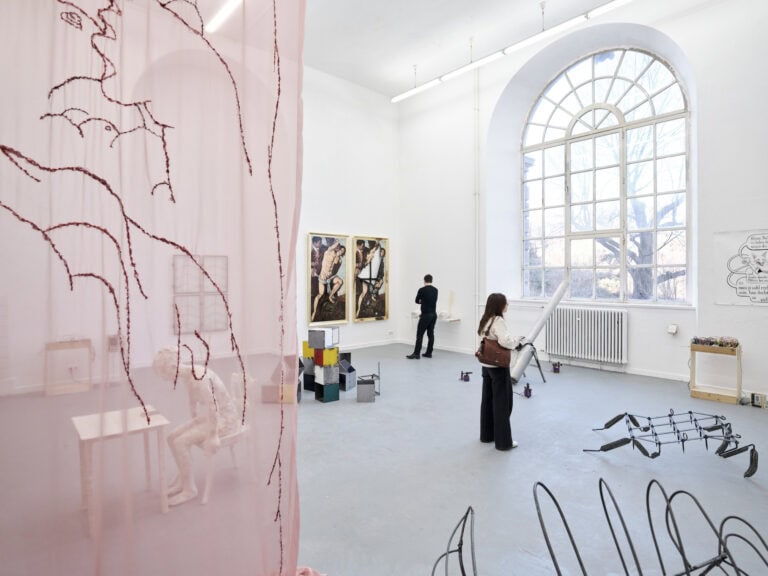 Foto: Ansicht eines Raumes mit weißen Wänden, grauem Boden und einem großen Bogenfenster. Im Raum und an den Wänden befinden sich eine Vielzahl von Objekten, Textilien und Malereien. Im Raum befinden sich Menschen.