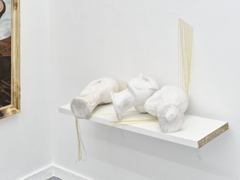Foto: Nahaufnahme von weißen, mit Schnüren versehenen Objekten auf einem weißen Regalbrett vor einer weißen Wand.