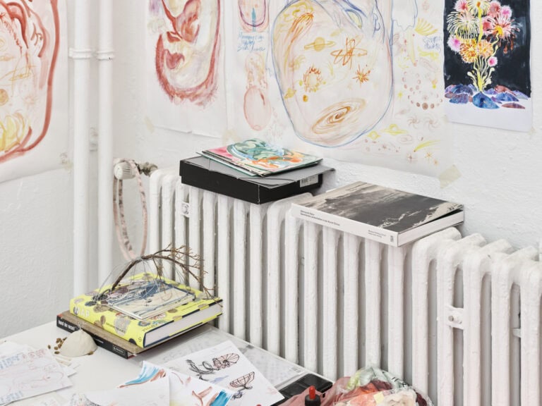 Foto: Nahaufnahme von Objekten, Büchern und Papierarbeiten, die sich auf einer weiß gestrichenen Wand, einer weiß gestrichenen Heizung und einem weißen Tisch befinden.