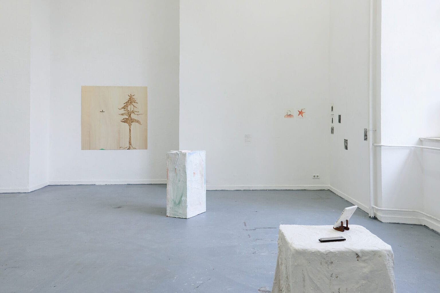 Foto: Ein Raum mit weißen Wände und grauem Boden. Im Raum zwei Objekte. An der Wand ein flaches Objekt im Mittelfromat und eine Gruppe von Papierarbeiten im Kleinformat.