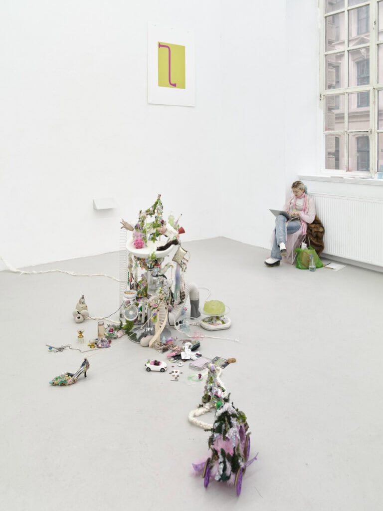 Foto: Ansicht eines Raumes mit weißen Wänden, gräulichem Boden und einem Fenster. Im Vordergrund das Bildes befinden sich bunte Objekte aus unterschiedlichen Materialien auf dem Boden. An der Wand befindet sich ein Papier mit farblichen Applikationen. Vor dem Fenster sitzt ein Mensch auf einem Stuhl und hält einen Computer auf dem Schoß.