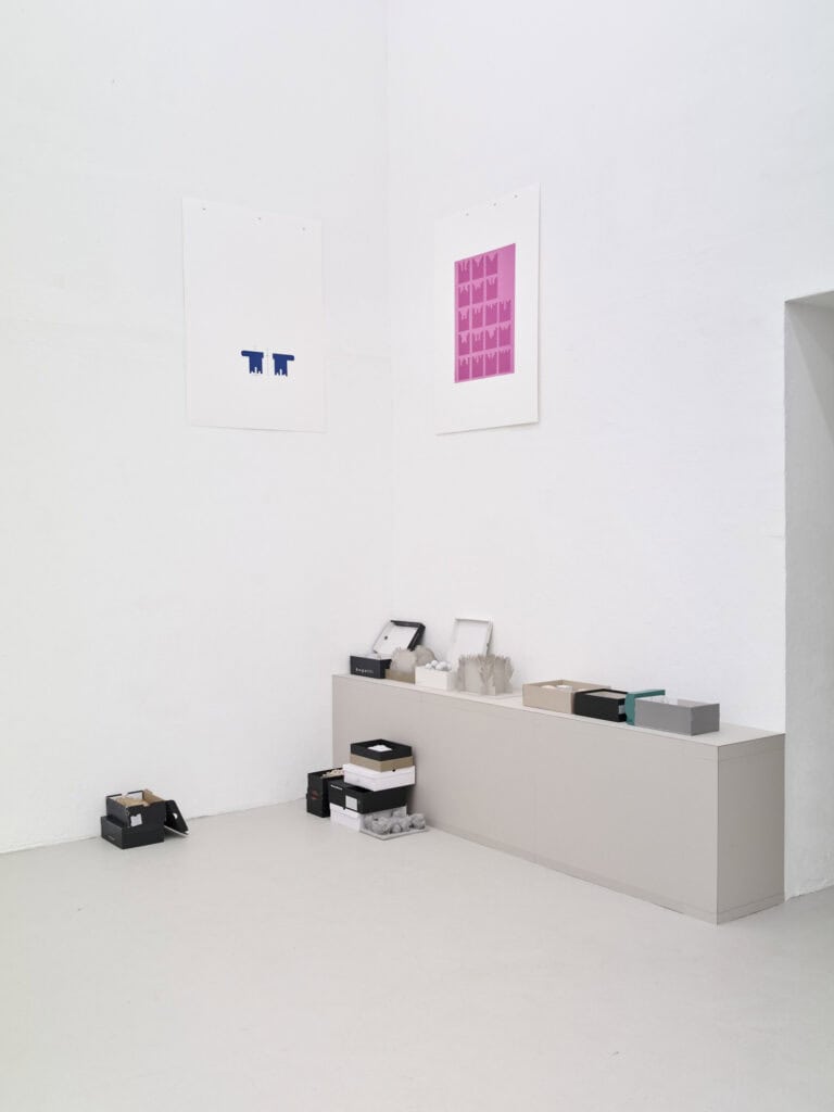 Foto: Ansicht eines Raumes mit weißen Wänden, grauem Boden und einem wandnahen, grauen Podest. An der Wand befinden sich Papiere mit farbigen Applikationen. Auf dem Boden und auf dem Podest befinden sich Schachteln und Objekte, die zum teil geöffnet und zum teil geschlossen sind.