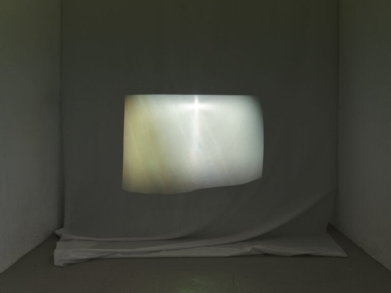 Foto: Ansicht eines abgedunkelten Raumes mit weißen Wänden und grauem Boden. Eine der Wände ist abgehangen. Das Material wirft falten. Auf ihm befindet sich ein unebenes, helles, verschiedenfarbiges Rechteck.