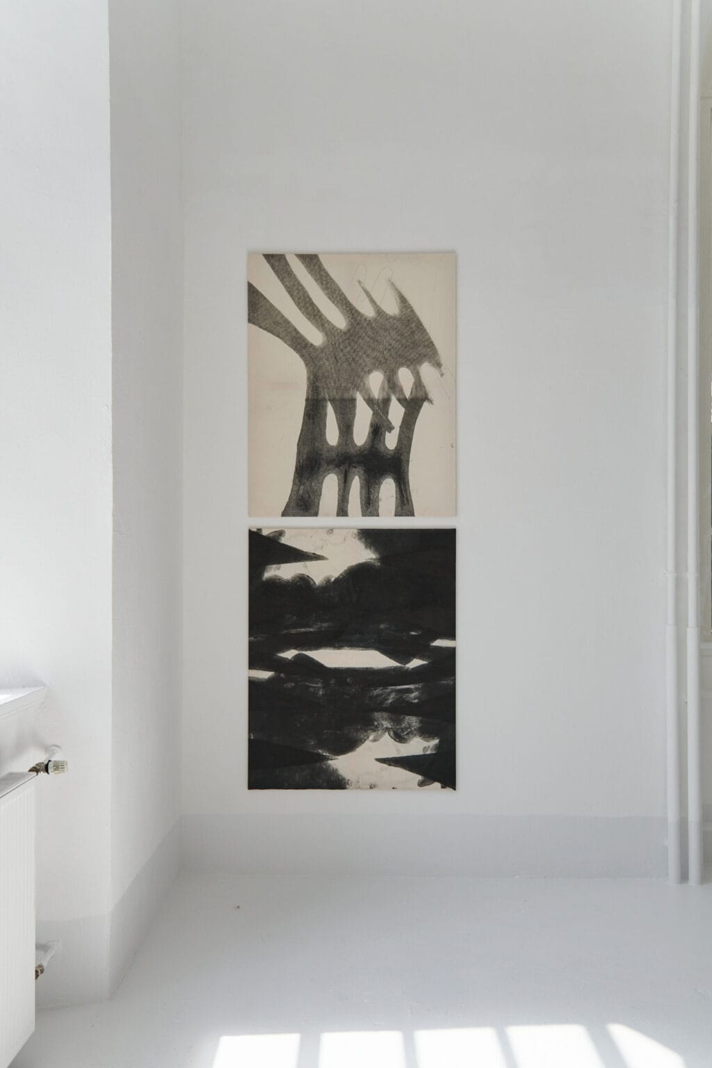 Foto: ein Raum mit weißen Wänden und hellgrauem Boden. An einer der Wände hängen zwei Leinwandarbeiten im gleichen Format, in einer Linie, untereinander. Durch den Raum bewegt sich ein Mensch.