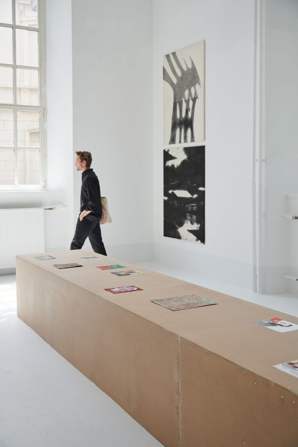 Foto: Ein Raum mit weißen Wänden, hellgrauem Boden und einem großen Fenster. Auf dem Boden steht ein länglicher, brauner Sockel auf dem kleinformatige Papierarbeiten liegen. An einer der Wände hängen zwei Leinwandarbeiten im gleichen Format, in einer Linie, untereinander. Durch den Raum bewegt sich ein Mensch.
