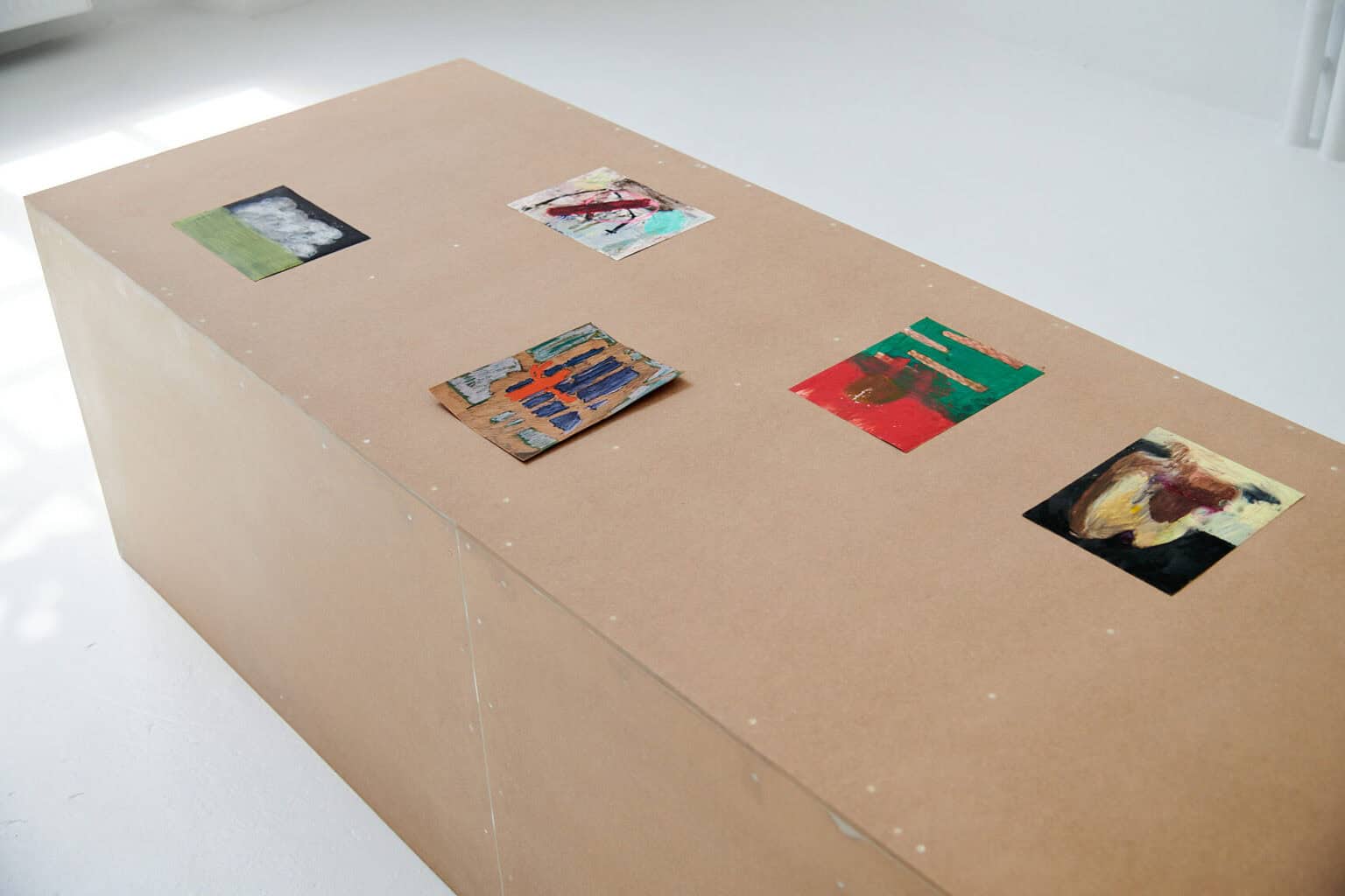 Foto: Ein länglicher, brauner Sockel auf dem fünf kleine Papierarbeiten liegen vor hellgrauem Boden.