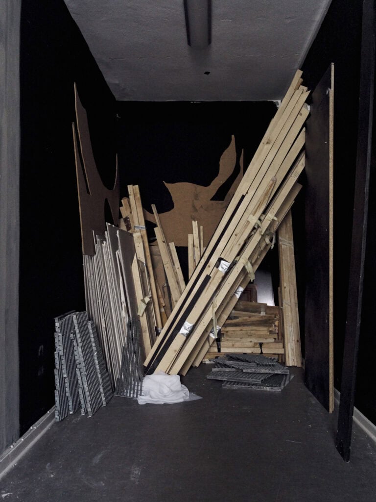 Foto: Ansicht eines schmalen Raumes mit schwarzen Wänden und grauen Boden, in dem unterschiedliche Materialien und Objekte eng aufeinander stapeln und aneinander liegen.