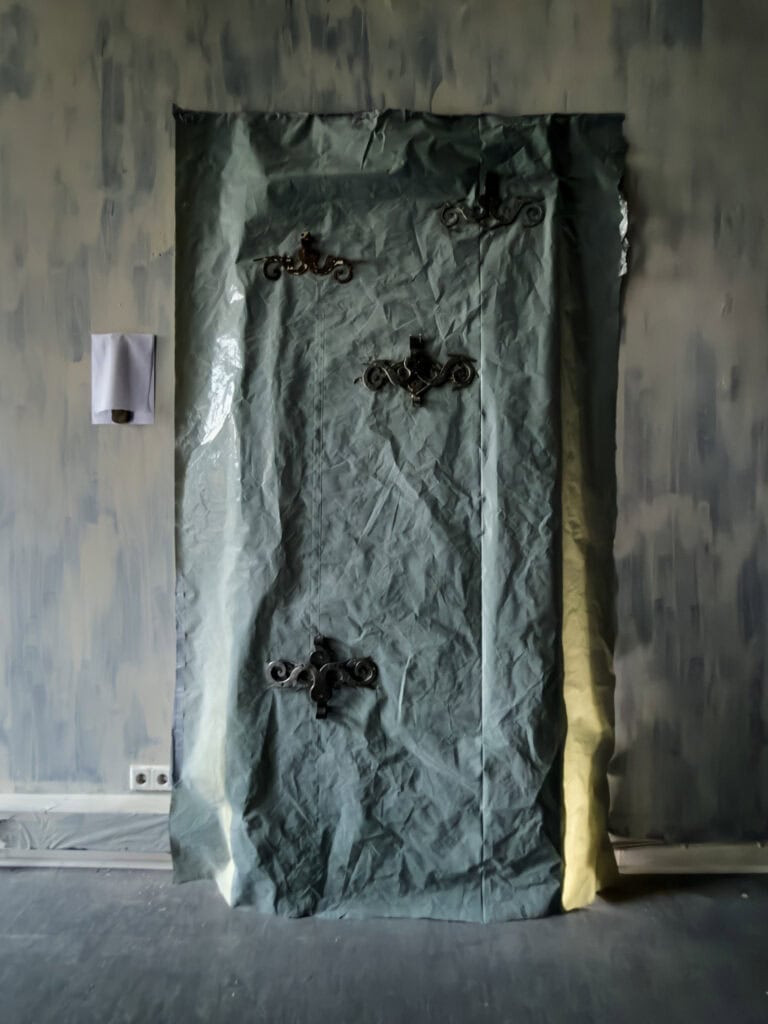 Foto: Installationsansicht eines hängenden Materials mit knittriger Oberfläche. Auf dem Material sind Objekte angebracht. Es hängt vor einer fleckig gestrichenen, cremefarbenen und blauen Wand. Der Boden ist gräulich.