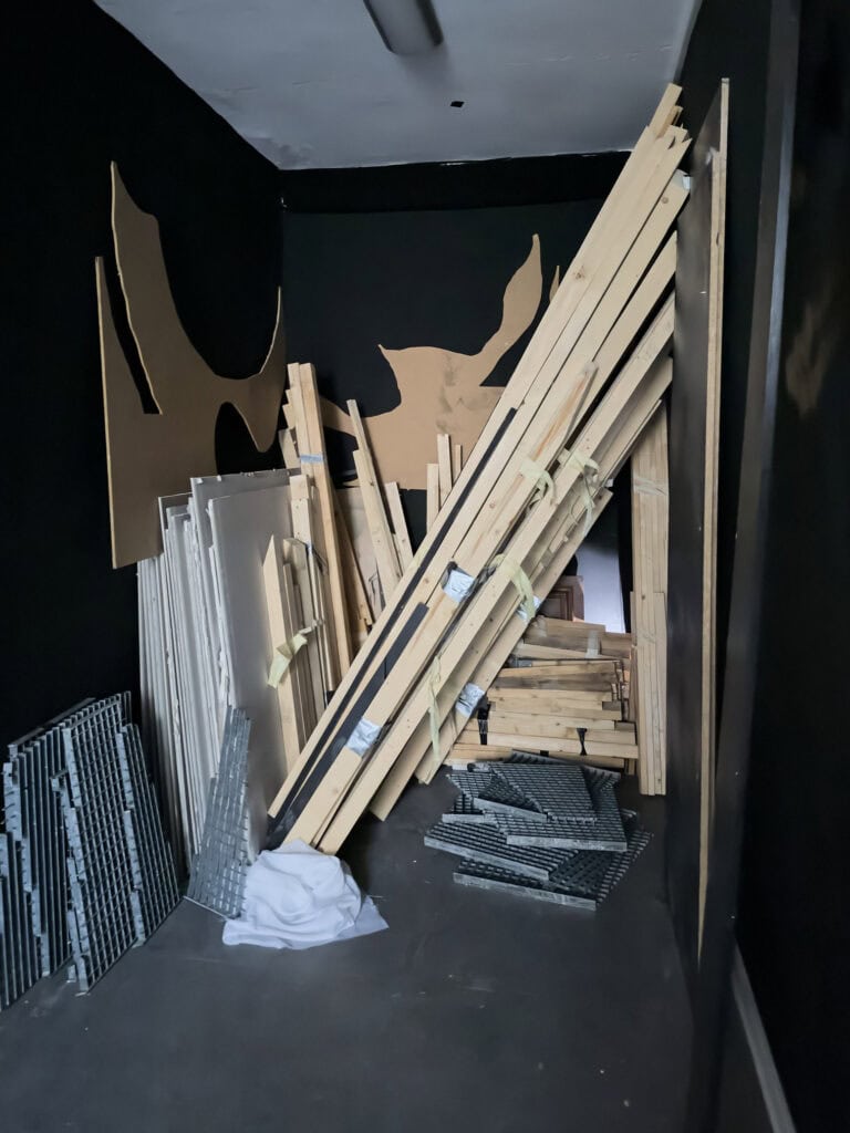 Foto: Ansicht eines schmalen Raumes mit schwarzen Wänden und grauen Boden, in dem unterschiedliche Materialien und Objekte eng aufeinander stapeln und aneinander liegen.