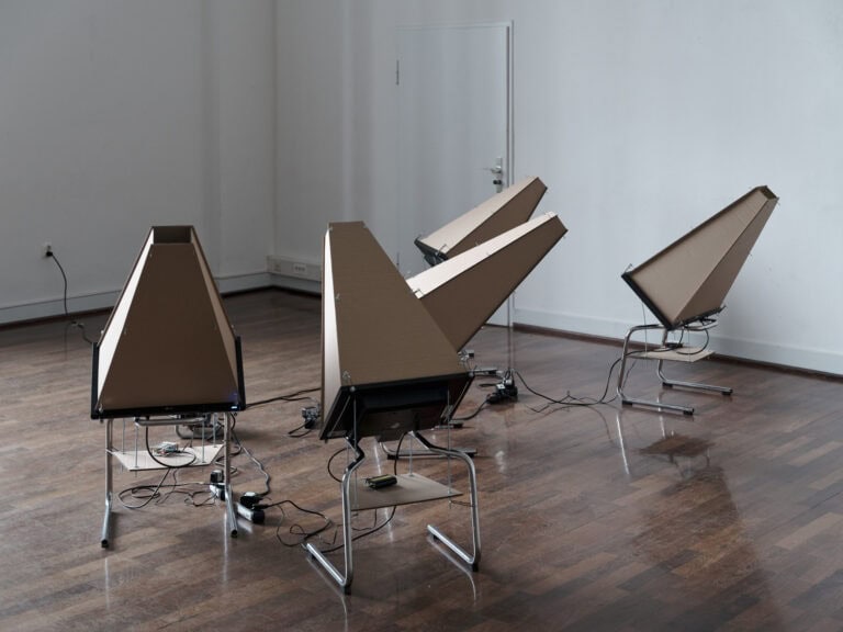 Foto: Installationsansicht von 5 ähnlich aussehenden Objekten, die an Kabel angeschlossen sind, in einem Raum mit weißen Wänden und braunem, reflektierendem Boden.