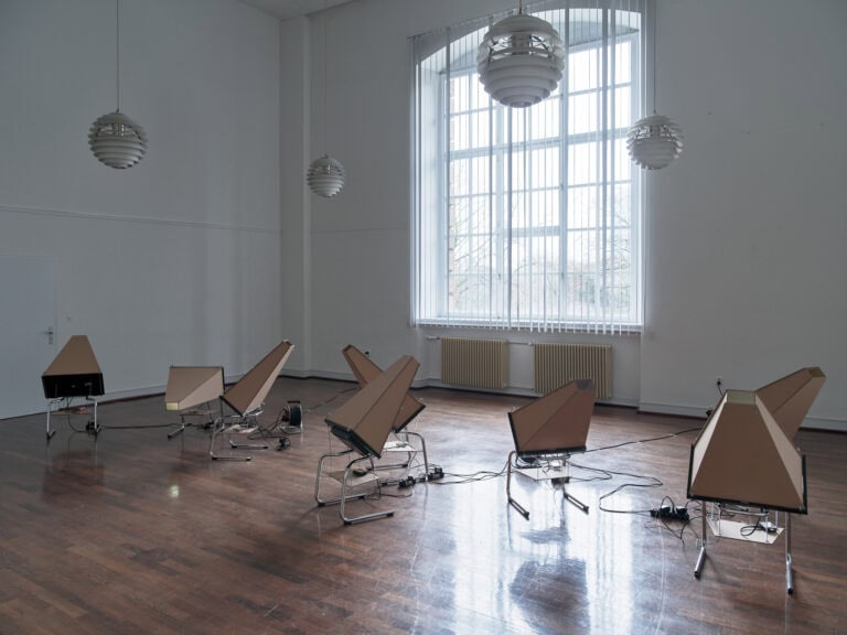 Foto: Ansicht eines Raumes mit weißen Wänden, Lamellen-Gardienen und mit braunem, spiegelnden Boden. Im Raum befinden sich acht ähnlich aussehende Objekte, die an Kabel angeschlossen sind.