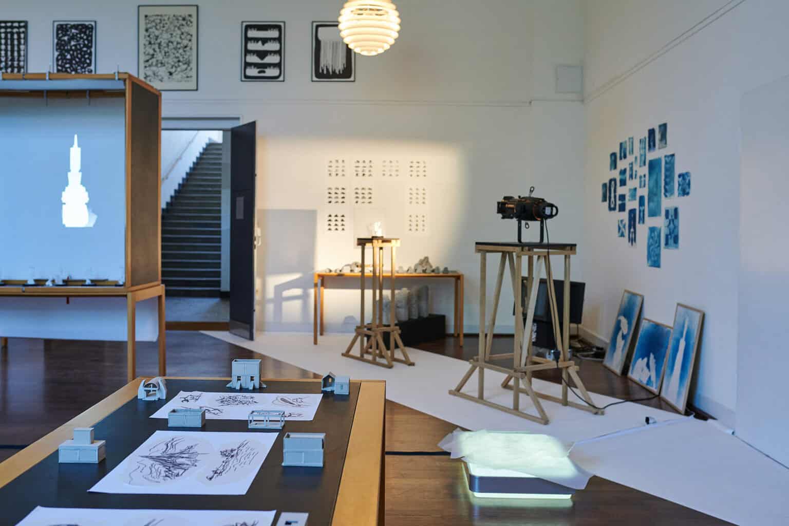 Foto: Ein Raum mit weißen Wänden und braunem Dielenboden. An den Wänden hängen und lehnen mehrere Gruppen von Objekten. Teile des Bodens sind in einer diagonalen Linie mit weißen Materialien bedeckt. Im Raum befinden sich mehrere Tische, Schaukästen und Konstruktionen, auf denen sich Objekte, Zettel und Technik befinden.