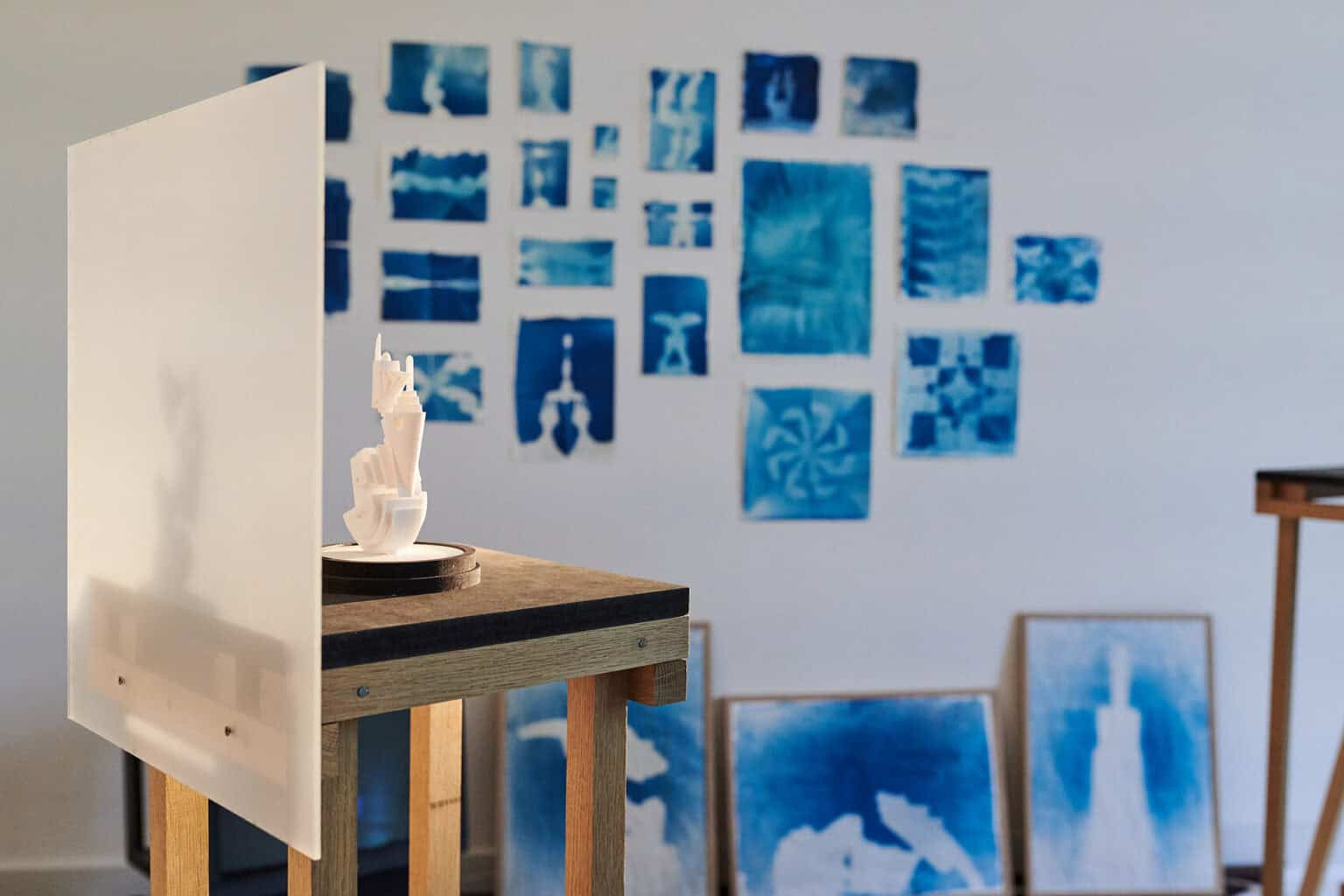 Foto: Ein Raum mit weißer Wand, an der viele Objekte hängen und lehnen. Im Raum ein Objekt aus unterschiedlichen Materialien, dass von Licht angestrahlt wird. Ein Schatten fällt auf eine in das Objekt integrierte Platte.