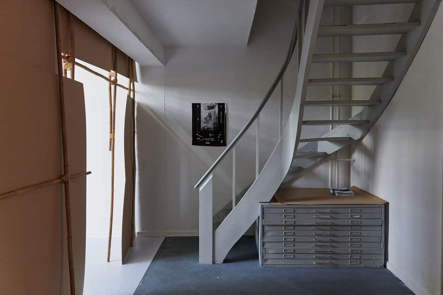Foto: Ein Raum mit grauer Wendeltreppe. An der Wand hängt ein flaches Objekt. Unter der Treppe steht ein grauer Zeichenschrank.