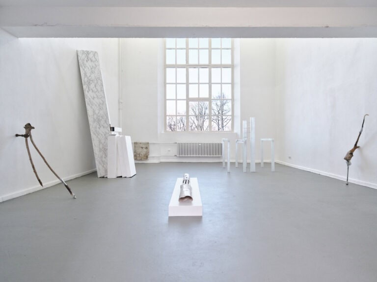 Foto: Ansicht eines Raumes mit weißen Wänden, grauem Boden und einem großen Fenster. Im Raum befinden sich mehrere Objekte, die an den Wänden lehnen, auf Sockeln liegen oder frei im Raum stehen.