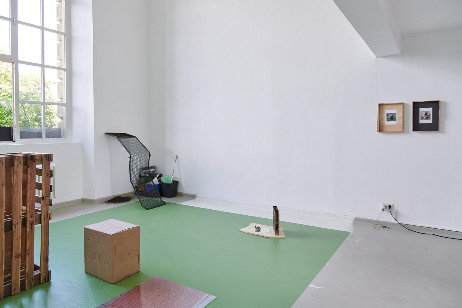 Foto: Ein Raum mit weißen Wänden und einem großen Fenster. Der Boden ist in der linken hälfte des Raumes, bis auf einen Streifen von mehreren Zentimetern entlang der Wände, grün gestrichen. Der Randstreifen ist grau. der rechte Teil des Raumes ist einheitlich in einem anderen grau gestrichen. Auf dem Boden des Raumes stehen mehrere Objekte, manche von Ihnen lehnen an der Wand. An einer der Wände hängen, auf gleicher höhe, flache Objekte.