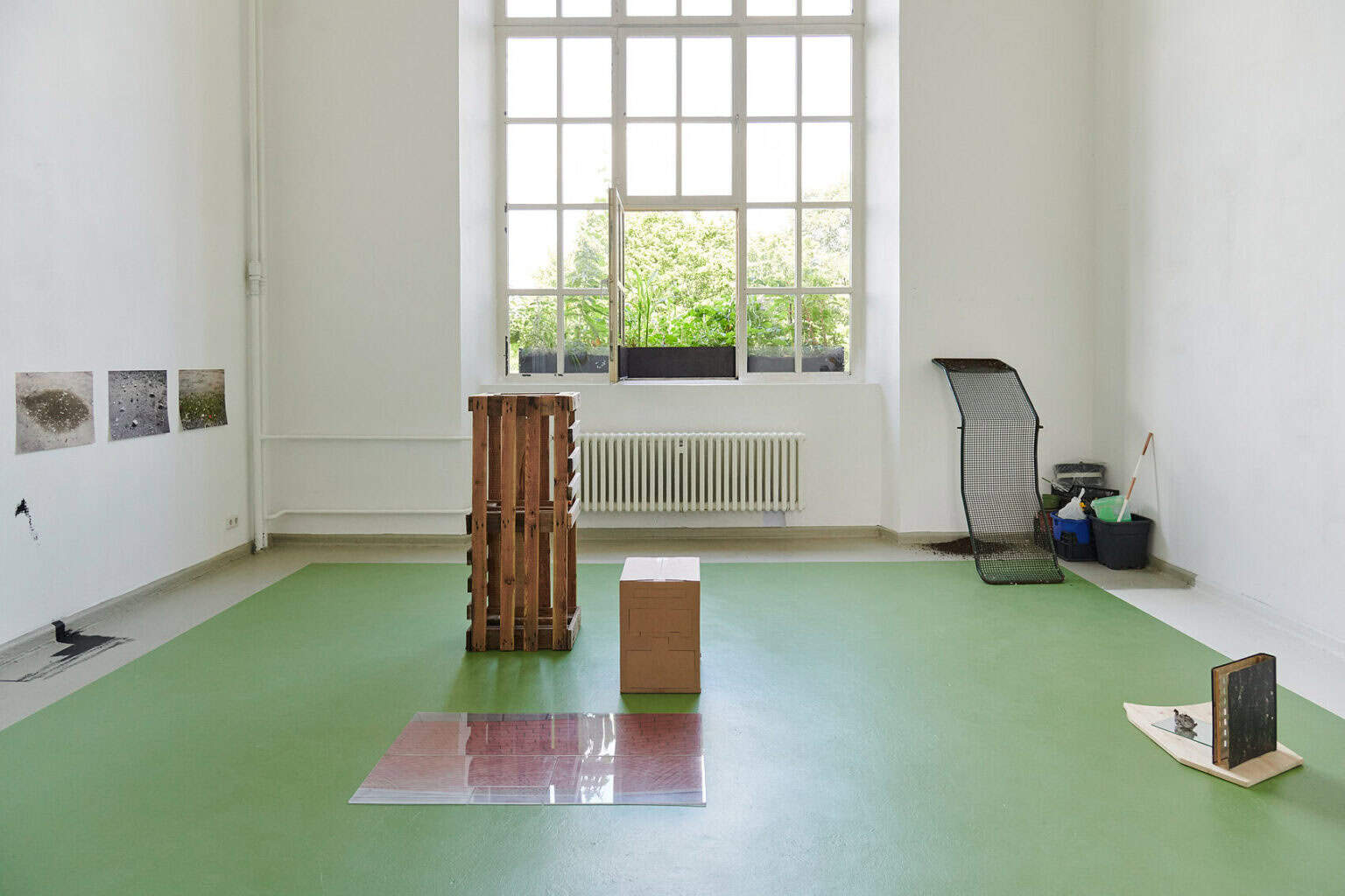 Foto: Ein Raum mit weißen Wänden und einem großen Fenster. Der Boden ist, bis auf einen Streifen von mehreren Zentimetern entlang der Wände, grün gestrichen. Der Randstreifen ist grau. Auf dem Boden des Raumes stehen mehrere Objekte, manche von Ihnen lehnen an der Wand. An einer der Wände hängen, auf gleicher höhe, flache Objekte. Unten an der Wand und auf dem drunterliegenden Boden befinden sich große Flecken aus schwarzer Farbe.