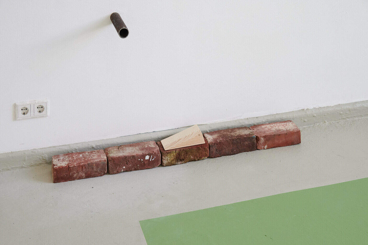 Foto: Detailaufnahme auf fünf Backsteinen, die in einer Reihe, an der Fußleiste des Raumes entlang, auf dem Boden liegen. Auf einem der Backsteine liegt ein Holzkeil. Teile des Bodens sind grau, andere Teile grün gestrichen. Aus der Wand ragt ein kurzes Metallrohr.
