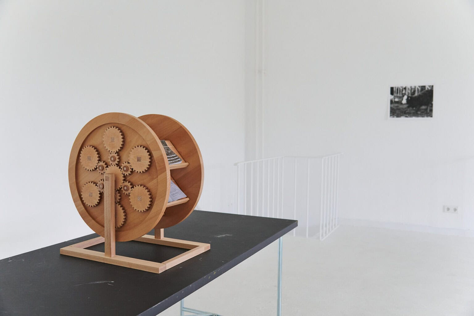 Foto: Ein reifenförmiges Objekt auf schwarzer Oberfläche. In dem Objekt liegen geöffnete und geschlossene Bücher und Hefte. An der Wand ein Objekt.