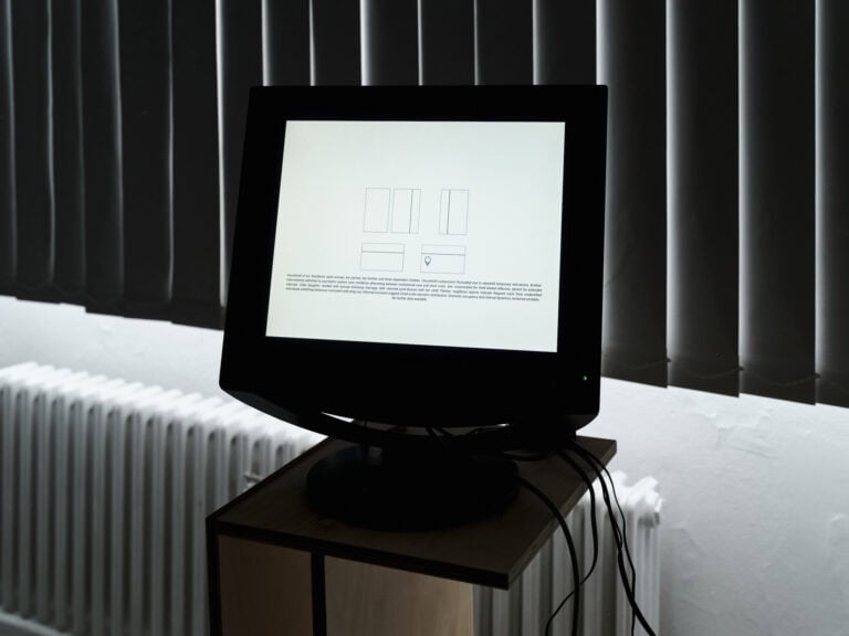 Foto: Nahaufnahme eines verkabelten Bildschirms, auf dem Abbildungen und Textelementen zu erkennen sind. Im Hintergrund verschwommen Lamellen-Gardienen und ein Heizkörper.