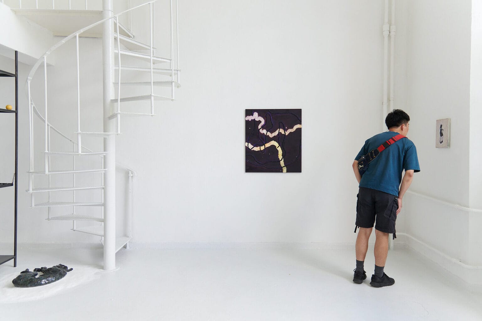 Foto:Ein Raum mit weißen Wänden, weißem Boden und weißer Wendeltreppe. Auf dem Boden liegt ein glänzendes Objekt auf einem Haufen weißem Sand auf dem Boden. An den Wänden hängen Arbeiten im Klein- und Mittelformat. Im Raum steht eine Person.