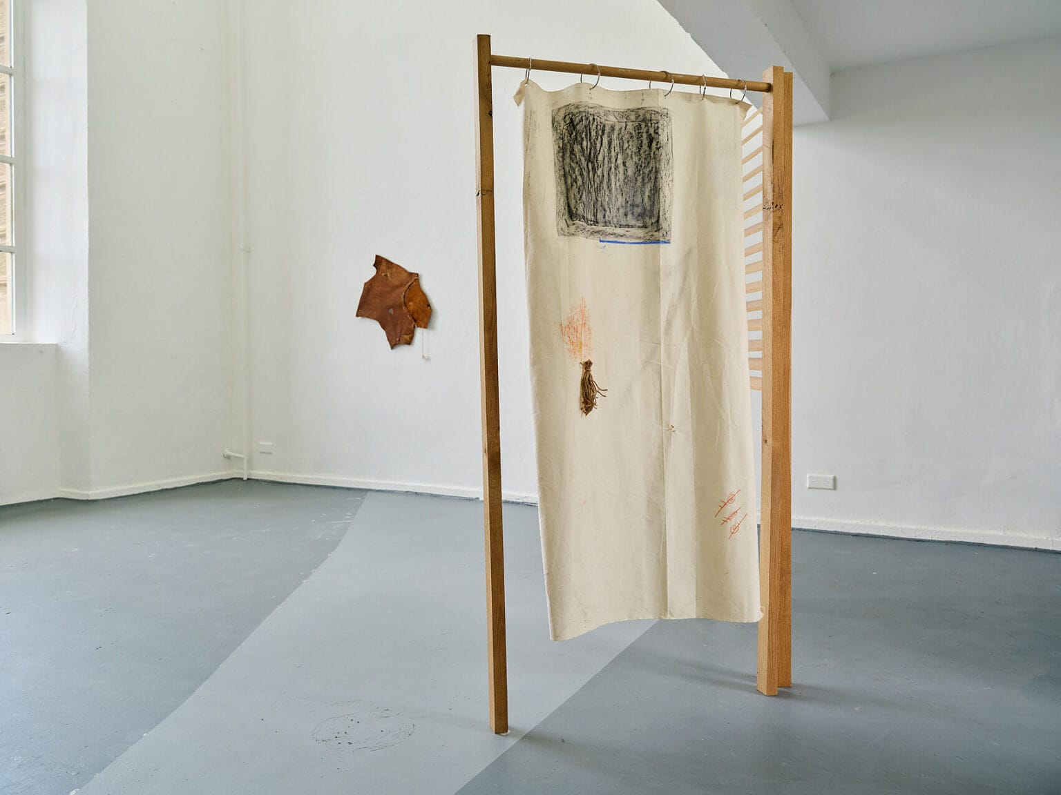 Foto: Ein Raum mit weißen Wänden, grauem Boden und einem Fenster. An der Wand hängen und im Raum stehen Objekte.