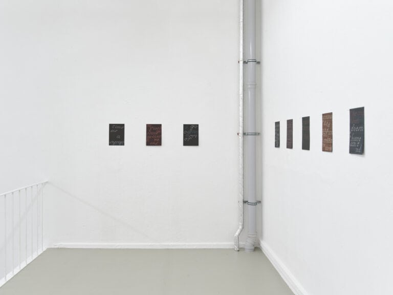 Foto: Ansicht eines Raumes mit weißen Wänden, Handlauf und grauen Rohren. An den Wänden hängen acht kleinformatige Papierarbeiten, alle auf gleicher Höhe.