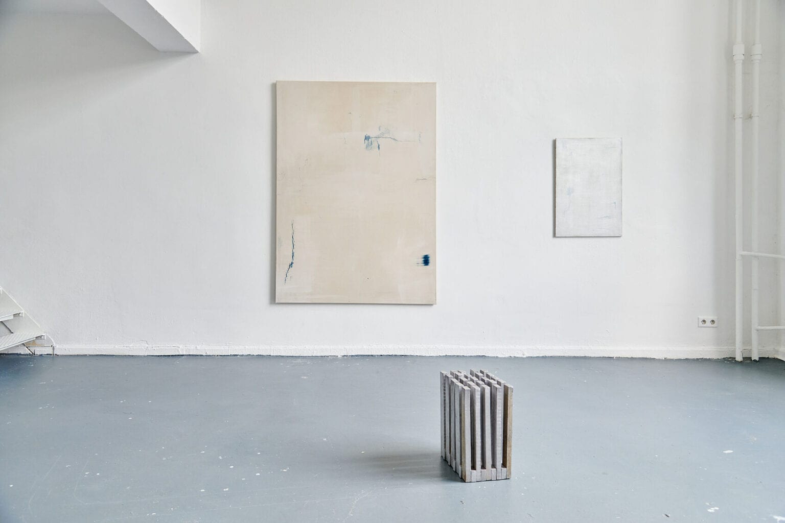 Foto: Ein Raum mit weißen Wänden und grauem Boden. An den Wänden hängen zwei Mittel- und Großformatige Arbeiten auf Leinwand. Auf dem Boden steht eine Skulptur.