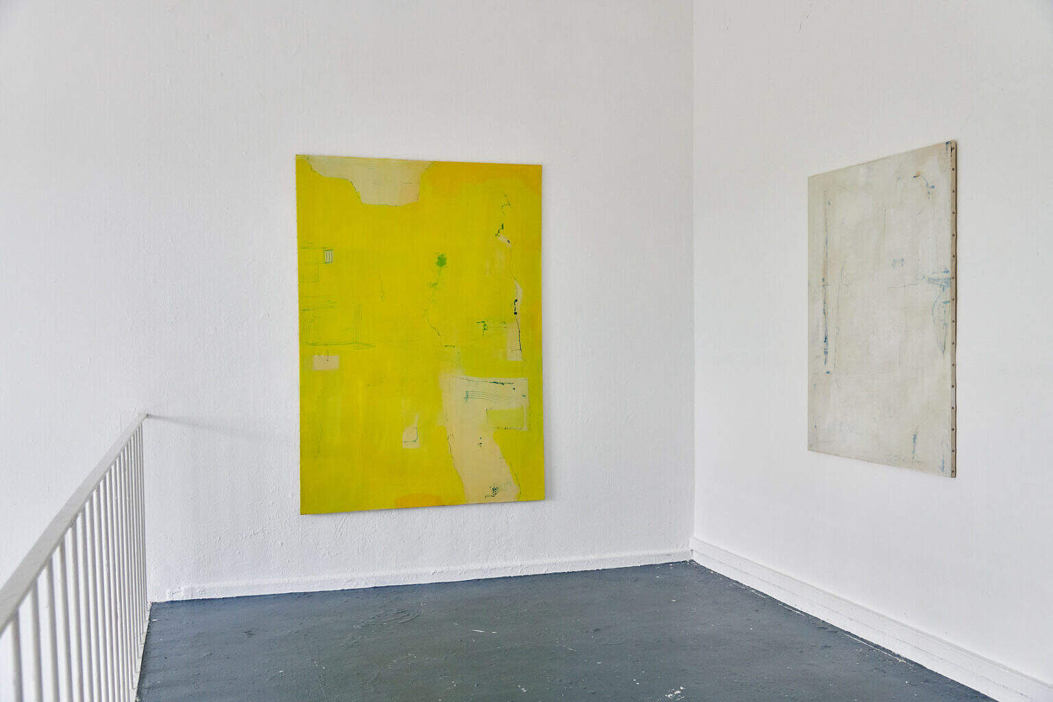 Foto: Die Raumecke eines Raume mit weißen Wänden, grauem Boden und Geländer. An den Wänden hängen zwei große, hochformatige Leinwände.