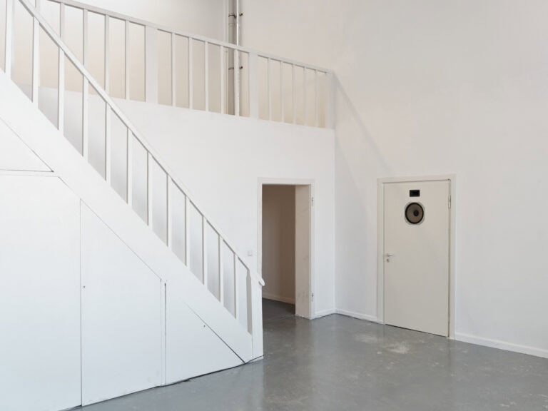 Foto: Ansicht eines Raumes mit einer durch eine Treppe erreichbaren, zweite Ebene. Die Wände sind weiß gestrichen. Der Raum ist leer. In einer im Raum befindlichen, ebenfalls hell gestrichenen Tür, sind zwei Objekte eingelassen.