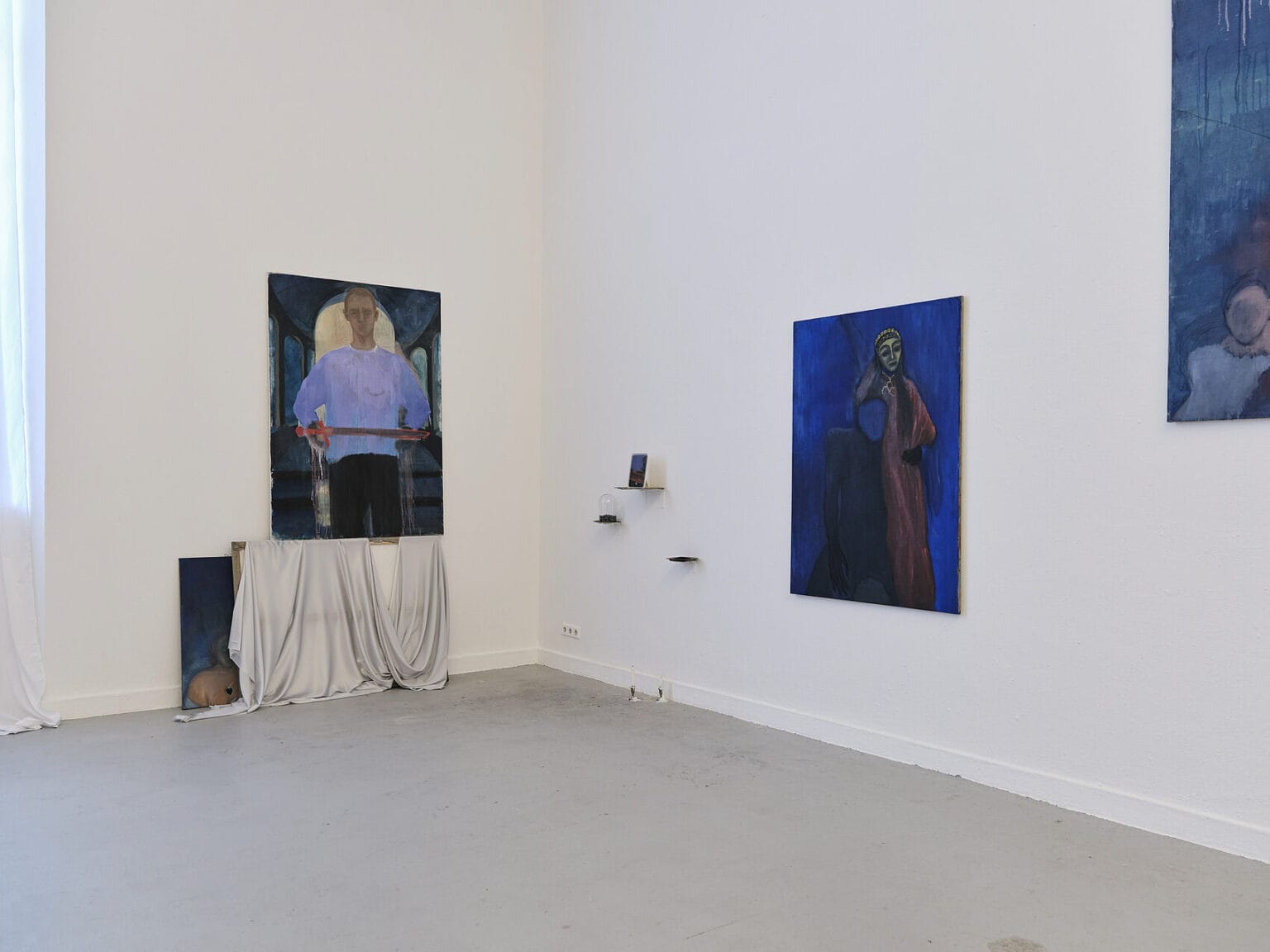 Foto: Raum mit weißen Wänden und grauem Boden. An den Wänden hängen, auf unterschiedlicher Höhe, mittelformatige Leinwandarbeiten. unter und zwischen den Leinwänden befinden sich Objekte, die ebenfalls an der Wand hängen oder auf dem Boden stehen.