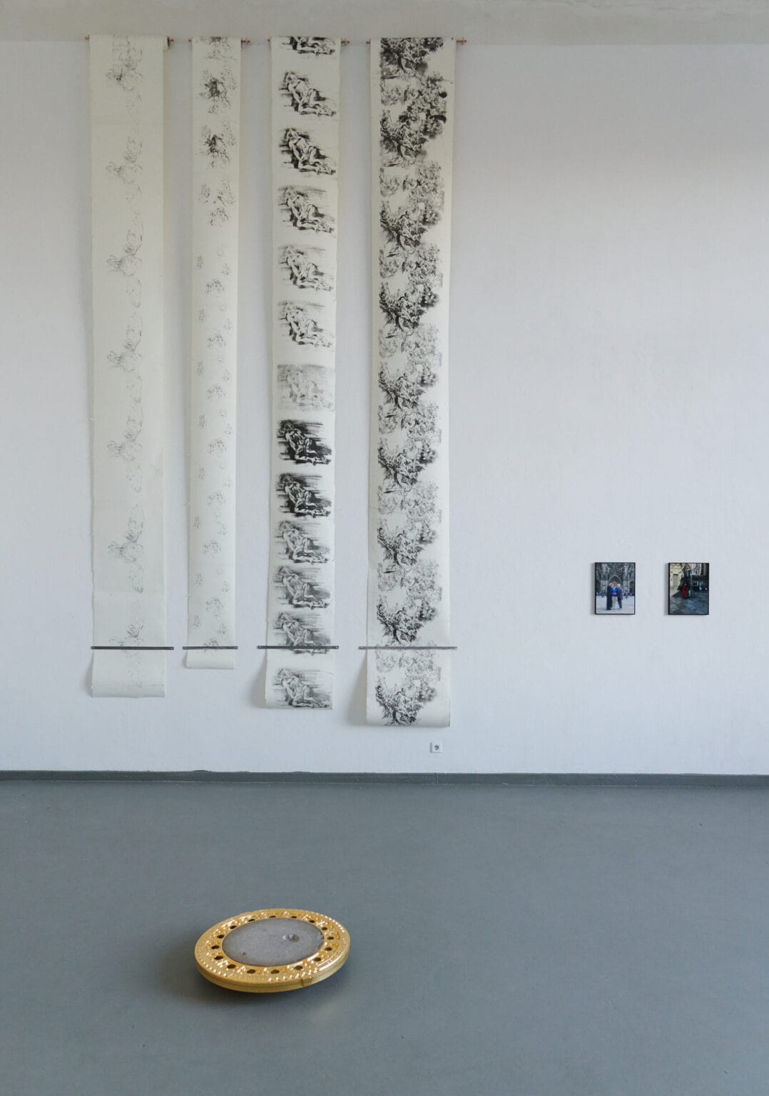 Foto: Ansicht eines Raume mit weißen Wänden und grauem Boden. An der Wand installiert sind vier bemalte Papierrollen, die vom oberen Teil der Wand bis kurz vorm Boden aufgerollt und am Ende mit einer Halterung befestigt sind. Daneben hängen zwei Objekte im gleichen Format. Auf dem Boden liegt ein weiteres Objekt.