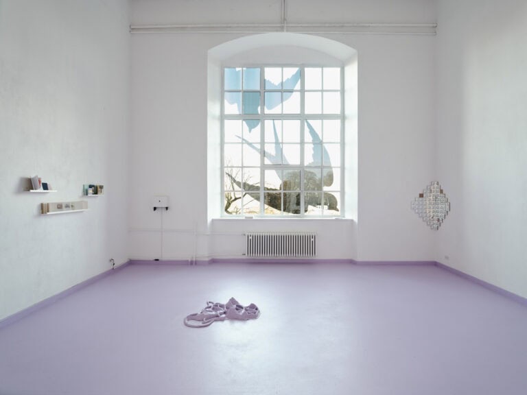 Foto: Raumansicht eines Raumes mit weißen Wänden, violettem Boden und einem großen Fenster. Die Scheiben sind mit Folien beklebt. An den Wänden befinden sich auf unterschiedlichen Höhen Objekte. Auf dem Boden lieht ein Objekt, dass die gleiche Farbe hat wie der Rest des Bodens.