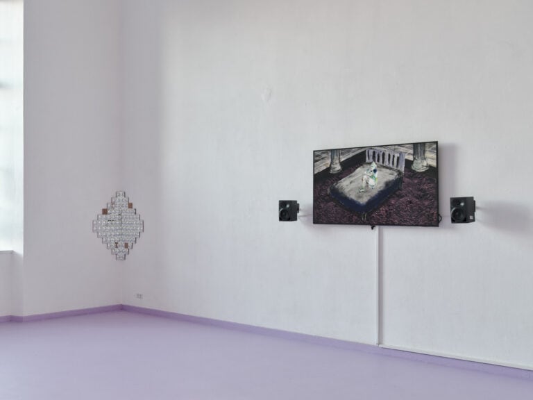 Foto: Aufnahme eines Raumes mit weißen Wänden und violettem Boden. An den Wänden befindet sich ein Objekt und ein Bildschirm, dass von zwei Lautsprechern eingerahmt ist.