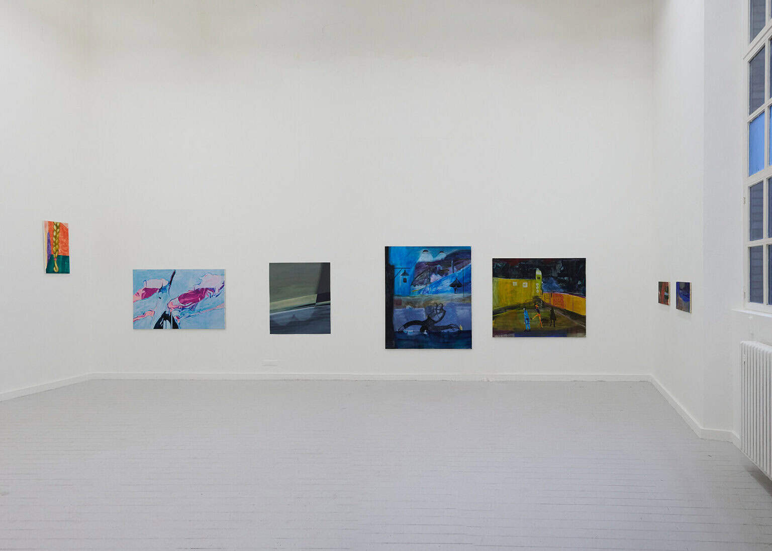 Foto: Ansicht eines Raumes mit weißen Wänden, grauem Dielenboden und einem großen Fenster. An den Wänden befinden sich Malereien im Klein-, Mittel- und Großformat. Manche von ihnen hängen in einer Reihe, auf ähnlicher Mittellinie, ein anderes deutlich höher.