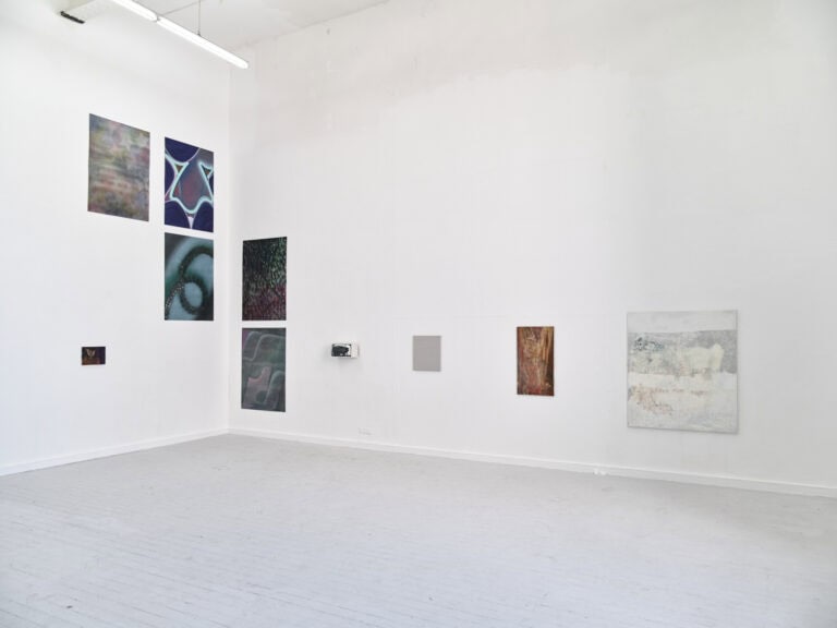 Foto: Aufnahme eiens Raumes mit weißen Wänden und grauem Dielenboden. An den Wänden hängen Malereien und Objekte.