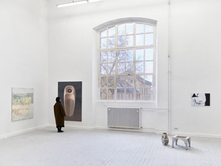 Foto: Aufnahme eines Raumes mit weißen Wänden, grauem Dielenboden und einem großen Bogenfenster. An den Wänden hängen Mittel- und Großformate. Auf dem Boden stehen Objekte. Vor einer der Malereien steht ein Mensch und betrachtet diese in Nahsicht.