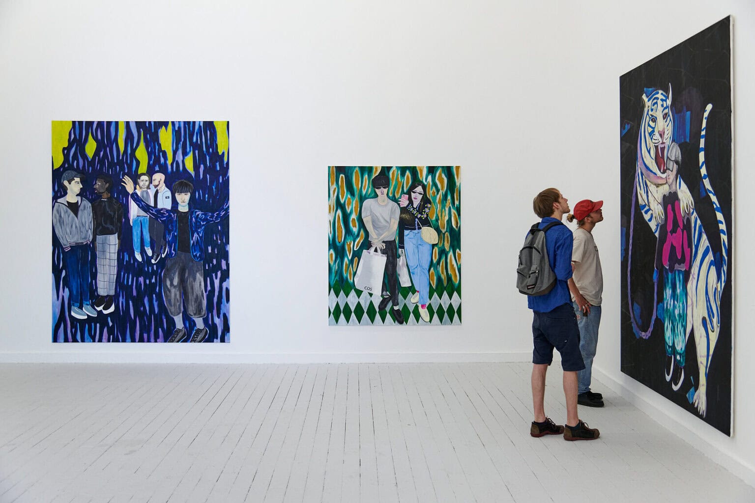 Foto: Ein Raum mit weißen Wänden und grauem Dielenboden. An den Wänden hängen großformatige Leinwände im Hochformat. Vor einem der Bilder stehen zwei Menschen.